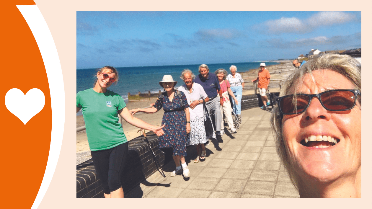 A photograph of an exercise class at the seaside. In bright sunshine, a line of older people hold onto the rail with one hand while doing exercises. In the foreground they are led by an instructor and the lady taking the photo is also in shot on the right, smiling and wearing sunglasses.