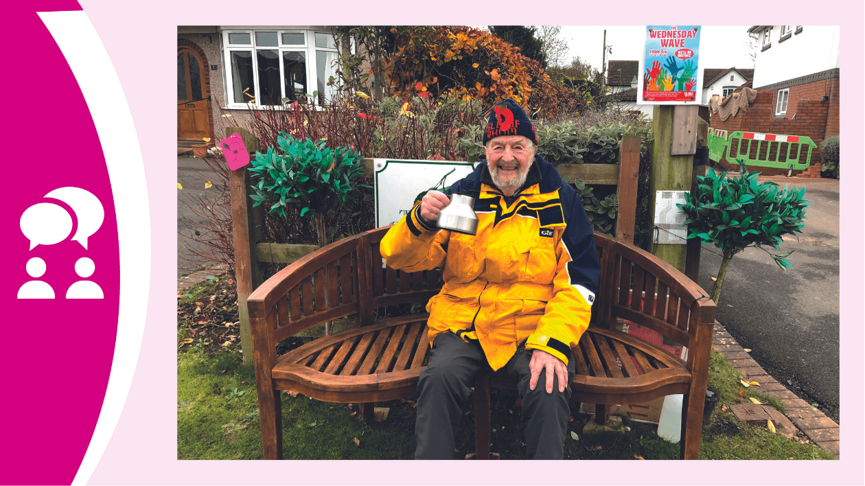 This is a photo of Roy sitting on his waving bench. He is wearing a hat, a bright yellow coat and holding up a mug (as if toasting the audience). He is sat on a wooden bench outside of his house.