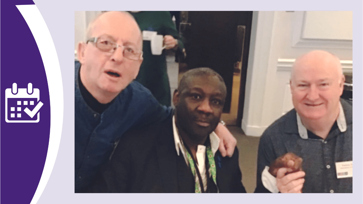 This photo is of the 3 men who wrote and performed the song 'I'm still climbing mountains' together. From left to right we have Paul Hitchmough (a white man wearing glasses and a navy shirt), Ronald Amanze (a black man wearing a white shirt, black jacket and sunflower lanyard), and Tommy Dunne (wearing a dark grey shirt). All three are looking towards the camera and are holding hands or have their arms around each other.