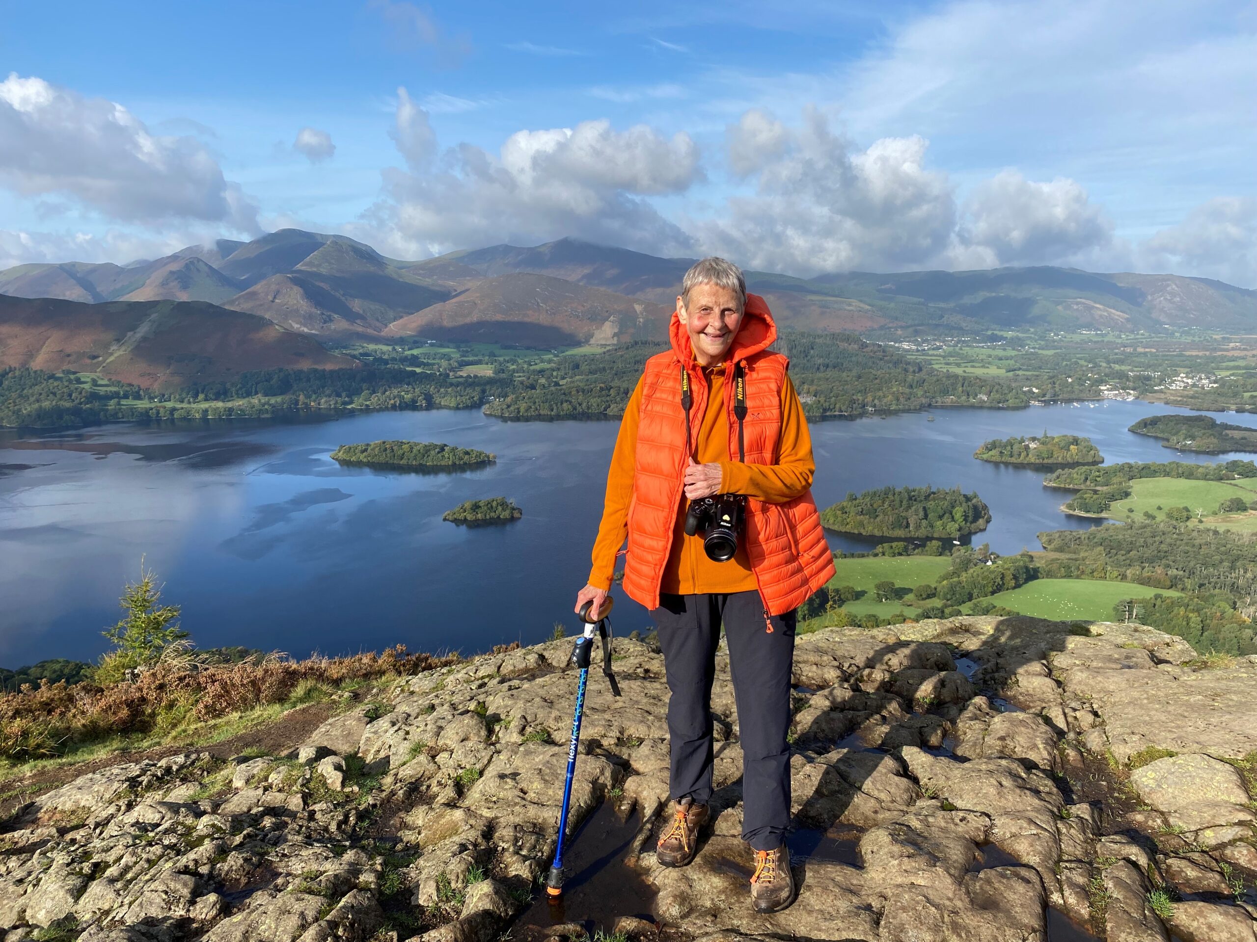 Wendy Mitchell in the foreground wearing walking boots, black trousers, a bright orange fleece and gilet. A professional-looking camera is around her neck and she has a bright blue walking stick. She is atop a mountain in the Lake District and behind her is a magnificent view of a large lake and, further in the background, another mountain range.