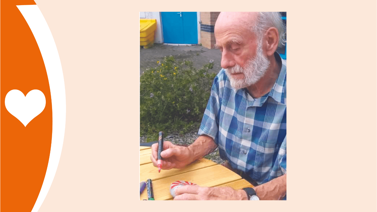 This photograph shows an older white haired gentleman with a beard. He is sitting outside in a blue checked shirt, using a red felt-tip pen to paint a pebble.