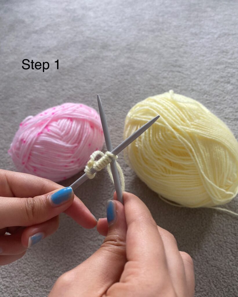 Two hands holding crossed knitting needles. The left needle is on top and has loops of wool over it. The right needle is crossed underneath. In the background we can see a pink ball of wool and a yellow ball of wool.