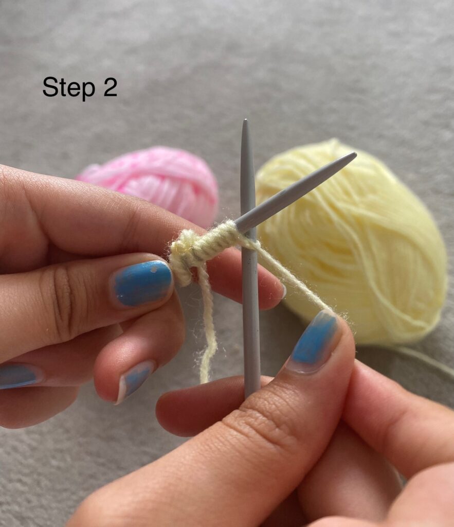 Two hands holding crossed knitting needles. The left needle is on top and has loops of wool over it. The right needle is hooked into the top loop from the left, with a thread of wool passing over the top of the loop.. In the background we can see a pink ball of wool and a yellow ball of wool.
