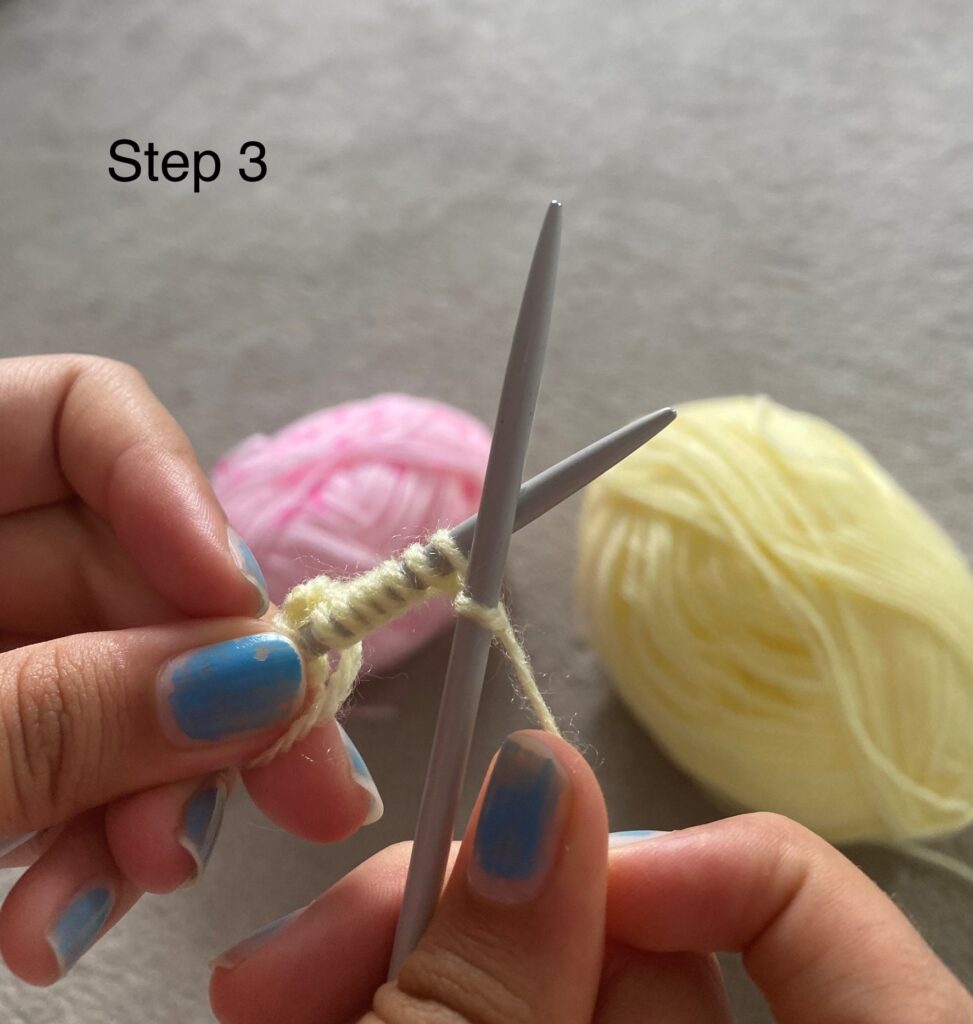 Two hands holding crossed knitting needles. The right needle is on top having made a stitch. There is a thread of wool behind the right needle, between the two needles. In the background we can see a pink ball of wool and a yellow ball of wool.