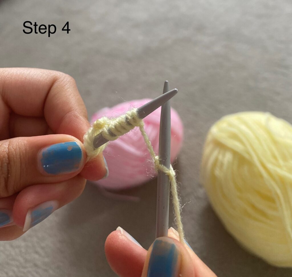 Two knitting needles at a small distance from one another. The left (on top) has several loops on the needle and the right (behind) has one loop. One thread links the two needles. In the background we can see a pink ball of wool and a yellow ball of wool.