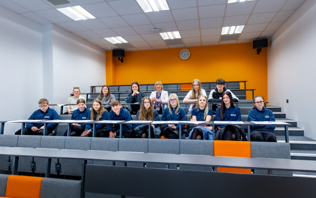 Year 10 and 12 Health and Social Care students from South Devon UTC. They are sitting in an orange and grey small lecture theatre with their teacher. Students are dressed in navy blue South Devon UTC sweatshirts.